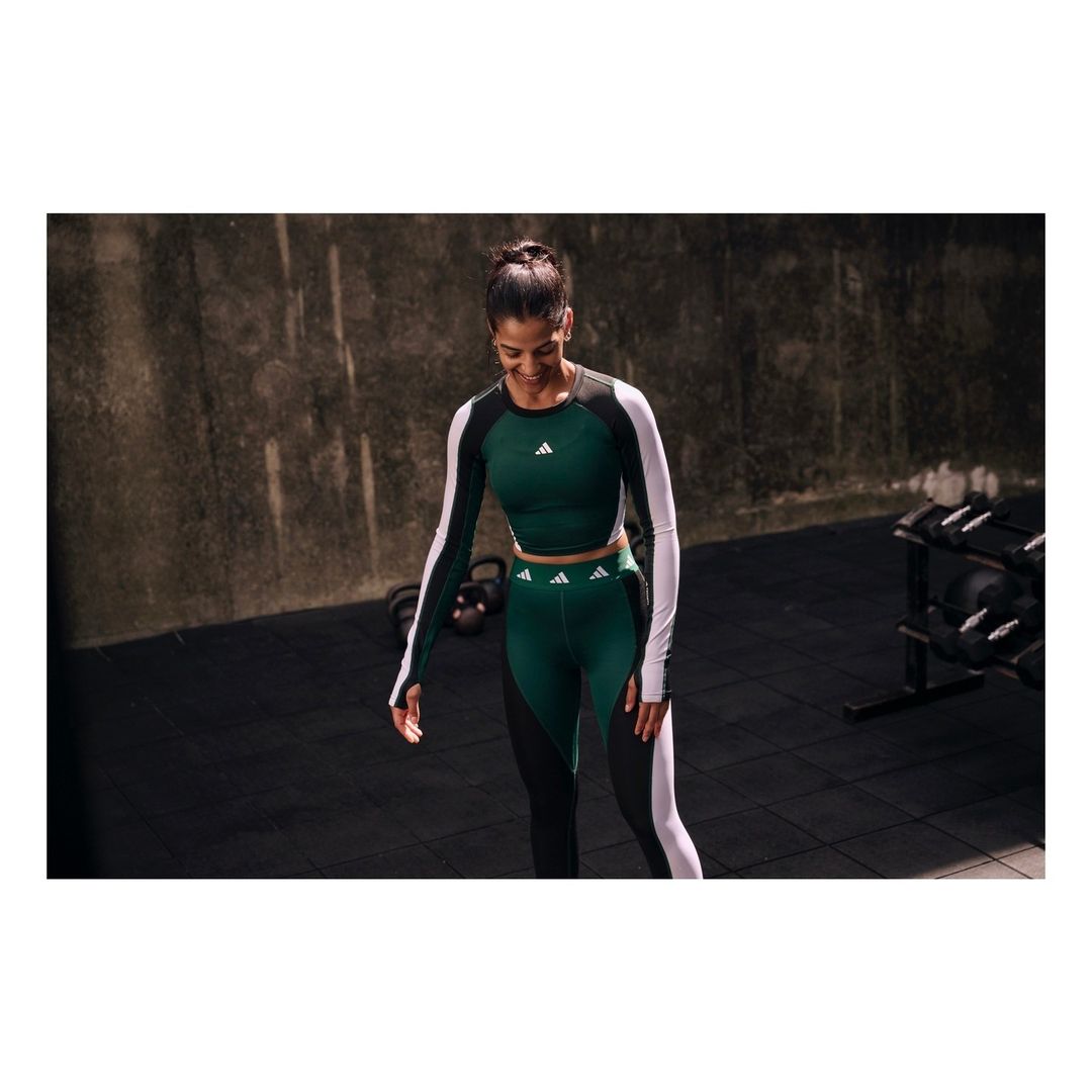 Athletic woman in a green and black color-blocked long-sleeve crop top and leggings smiles in a gym setting with weights nearby.