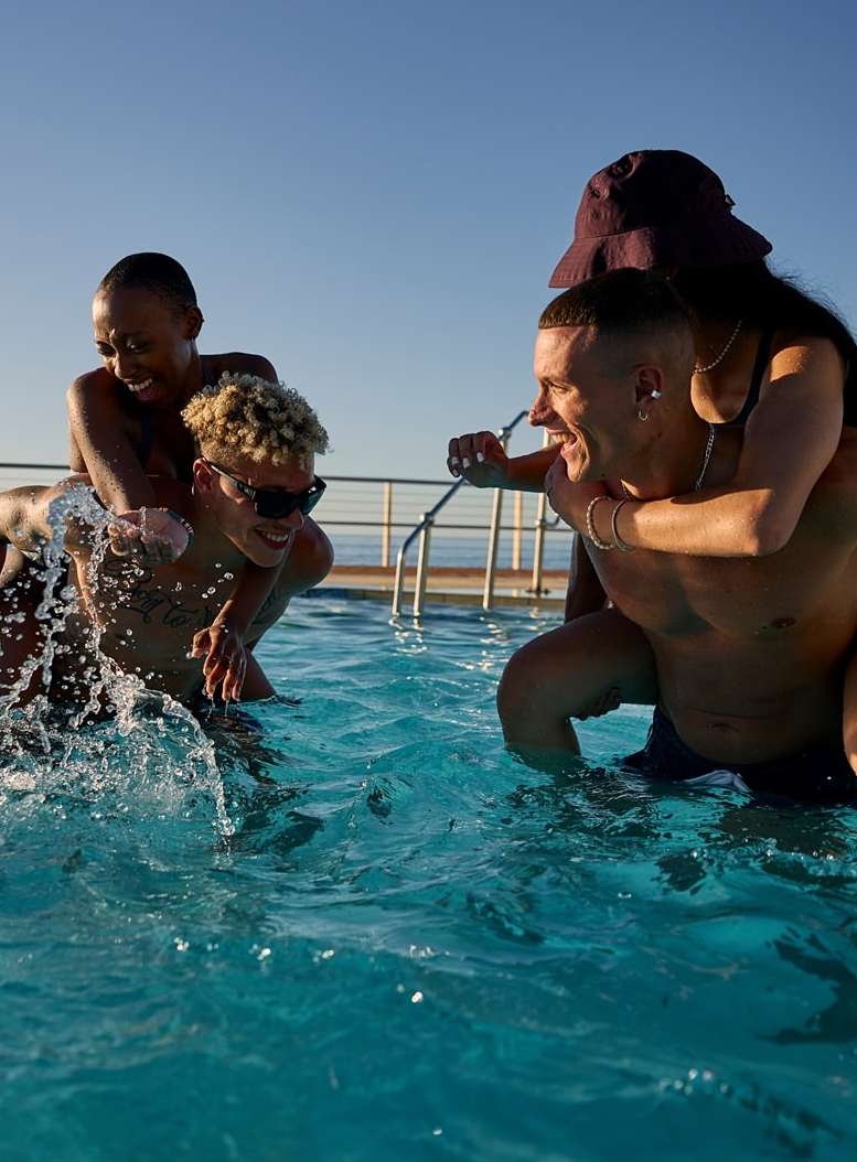 Four young adults, two men and two women, enjoy a sunny day in a pool, with two people giving piggyback rides, creating a fun and active scene.