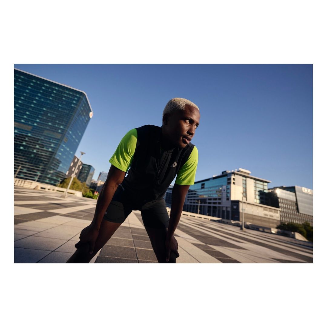 Athletic Black man resting with hands on knees after exercise. He's wearing a black vest, stands in a city plaza under a blue sky.