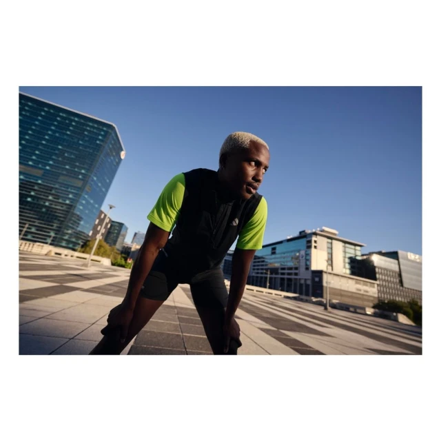 Athletic Black man resting with hands on knees after exercise. He's wearing a black vest, stands in a city plaza under a blue sky.