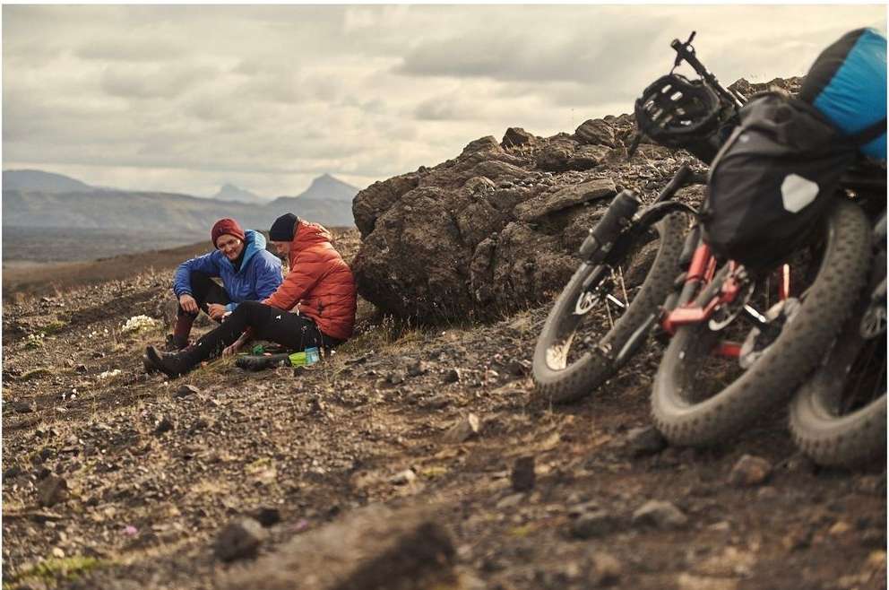 Two people in jackets sit on a rocky hill next to two fat tire bikes in a landscape setting with mountains under an overcast sky.