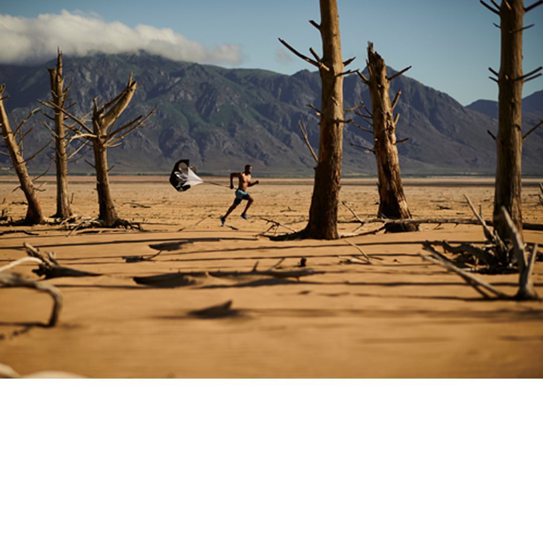 Adult male athlete runs across a tan, barren desert landscape, a parachute billowing behind, with distant mountains in the background.