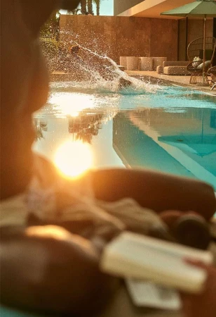 Backlit view over a lounging figure toward a person splashing into a bright swimming pool at sunset.