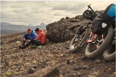 Two men resting near bicycles on a rocky terrain with mountains under an overcast sky, photographed by Florian Bison