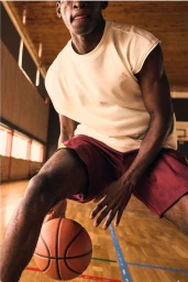 Close-up of a black man dribbling a basketball inside a gym, wearing an off-white t-shirt and maroon shorts.