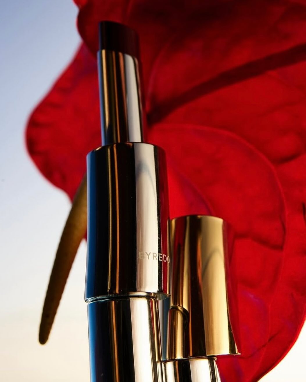 Close up of a metallic lipstick tube with its cap next to a large vibrant red anthurium flower against a pale sky.