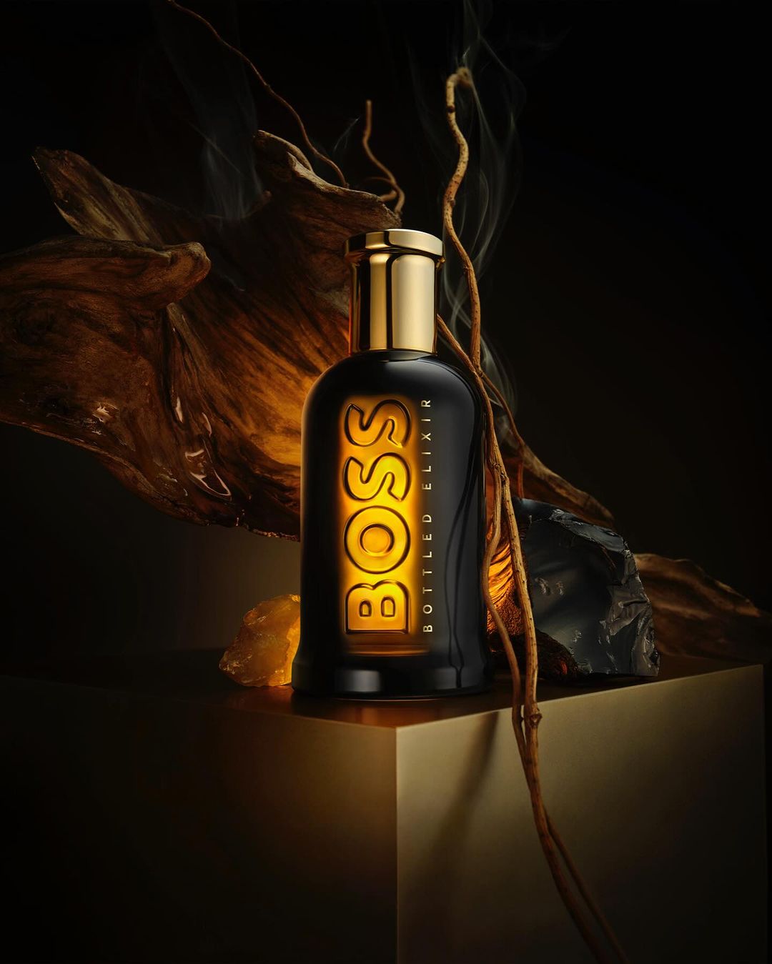A black BOSS Bottled Elixir perfume bottle with a golden cap and label on a golden geometric surface in a dark studio setting.