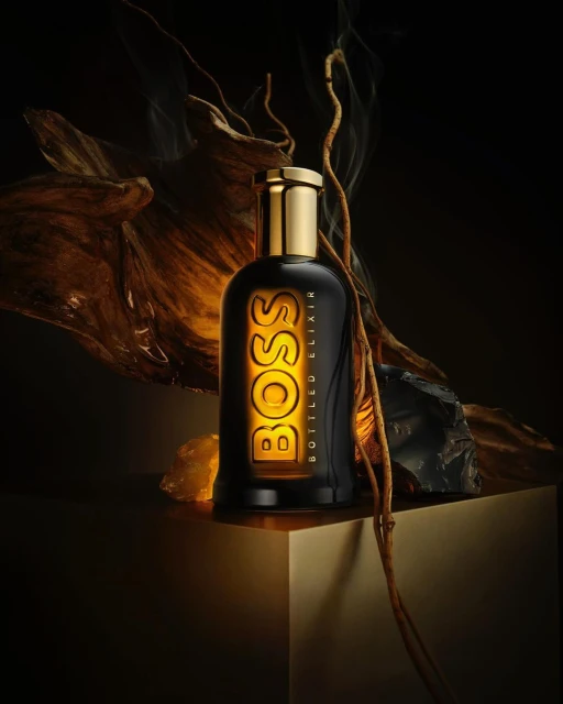 A black BOSS Bottled Elixir perfume bottle with a golden cap and label on a golden geometric surface in a dark studio setting.