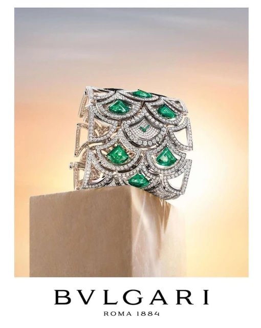 Elegant Bulgari bracelet, encrusted with diamonds and emeralds, showcased on a stone pedestal against a gradient sky backdrop.