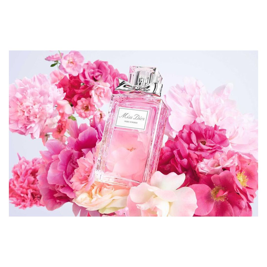 Elegant still life featuring a 'Miss Dior' perfume bottle with a silver bow, surrounded by vibrant pink peonies against a white backdrop.
