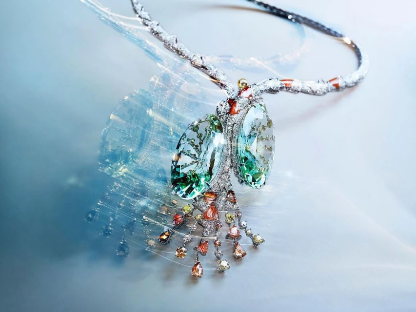 High-jewelry necklace featuring white gold and diamonds with green, orange, and yellow gemstones in a studio setting with light-blue background.