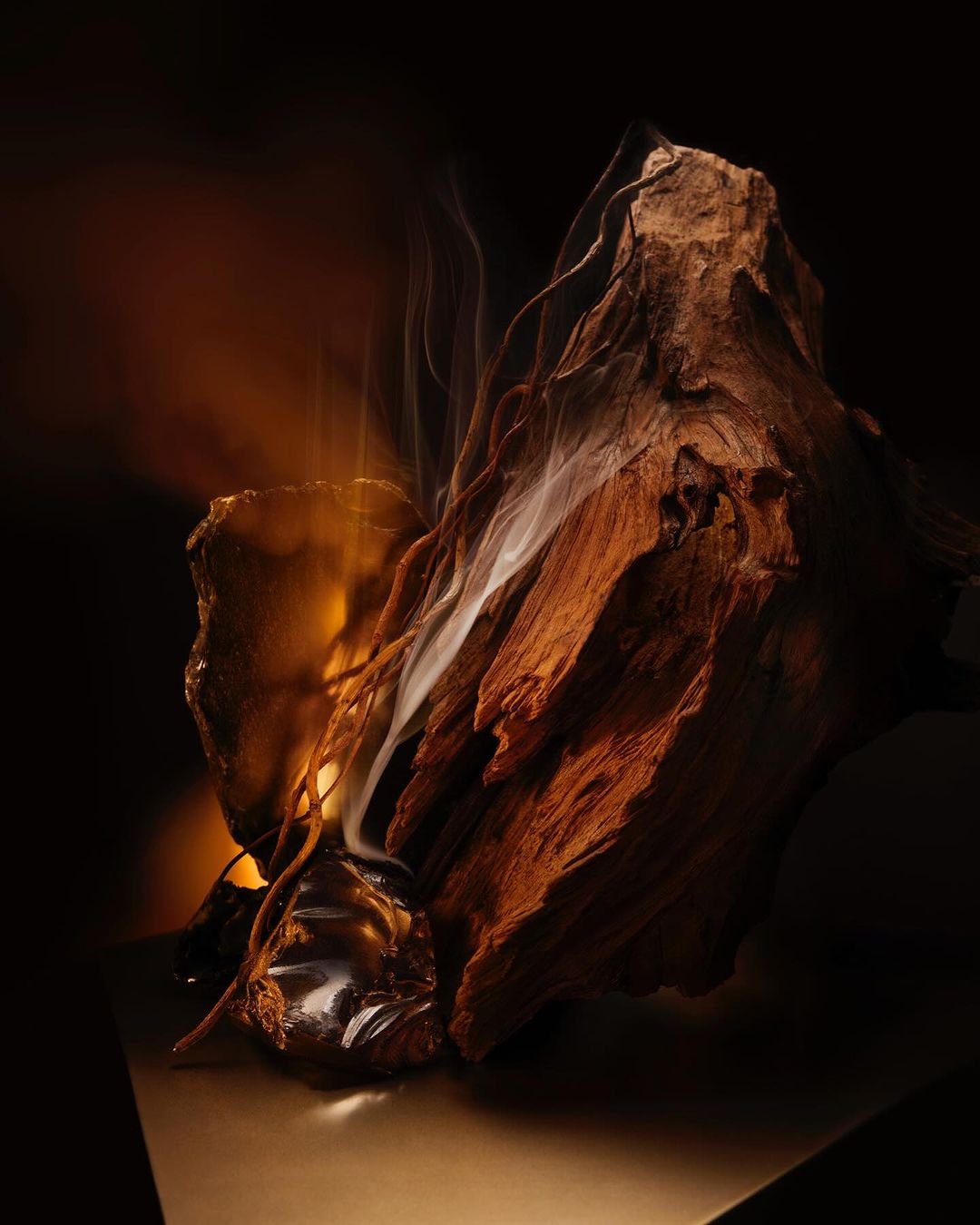 Dramatic still life shot featuring textured wood, amber resin, and ethereal smoke against a dark backdrop with warm lighting.