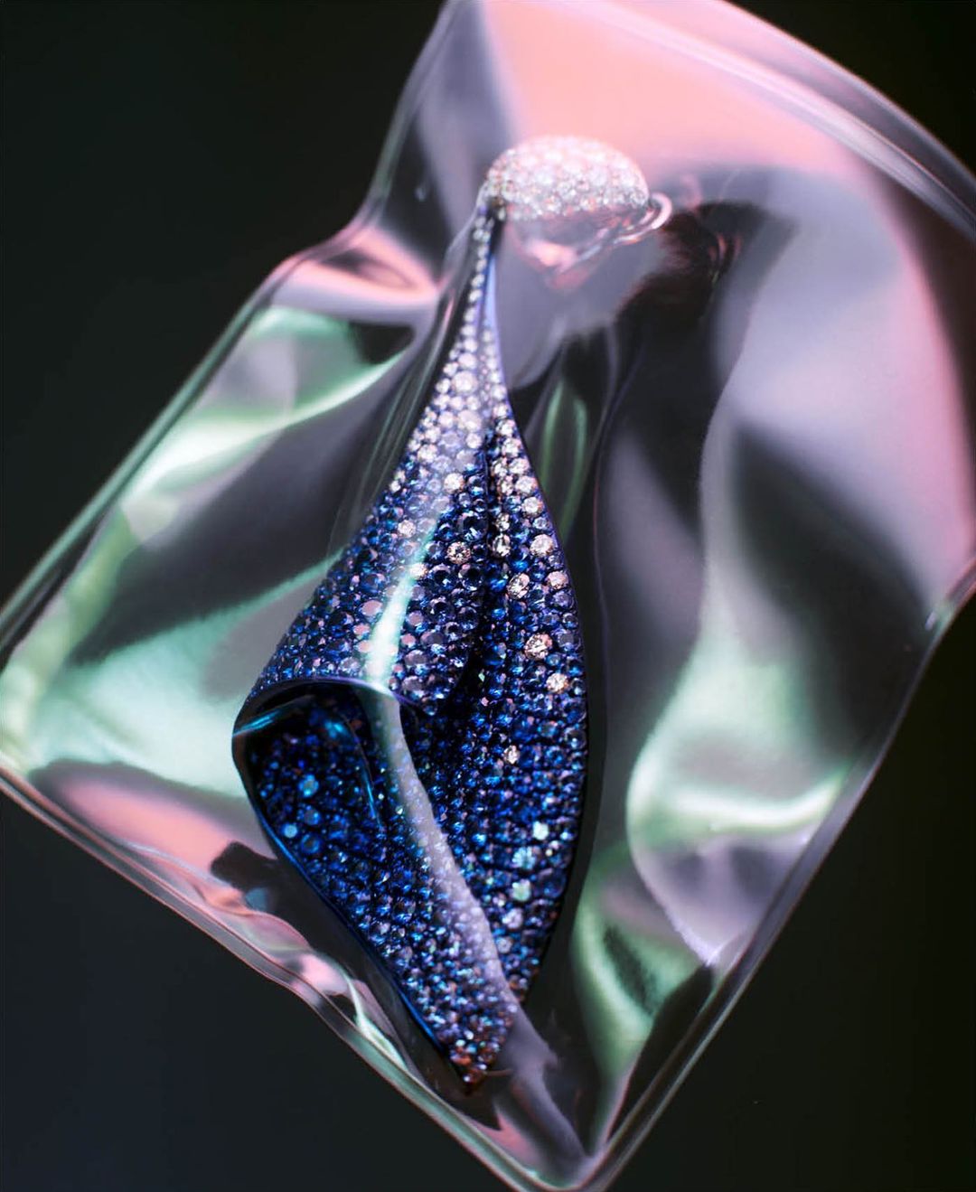 Elegant teardrop earring with blue and white gems displayed in translucent packaging with soft pink and green background lighting.