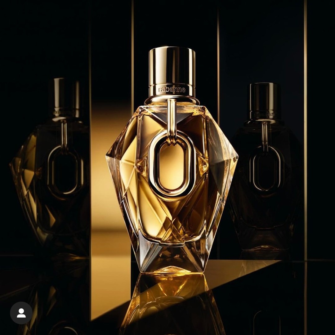 Three geometrically-faceted, golden-hued perfume bottles with metallic gold accents are arranged on a reflective gold surface against a dark backdrop.