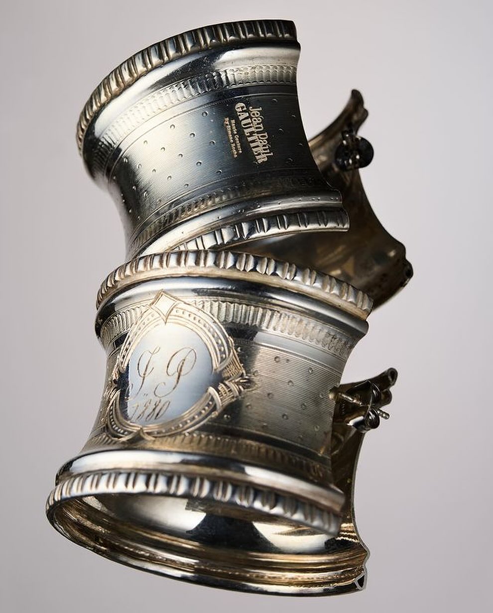 Silver napkin rings stacked show intricate designs and Jean Paul Gaultier branding, set against a soft gradient background.