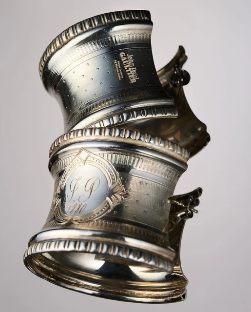 Silver napkin rings stacked show intricate designs and Jean Paul Gaultier branding, set against a soft gradient background.