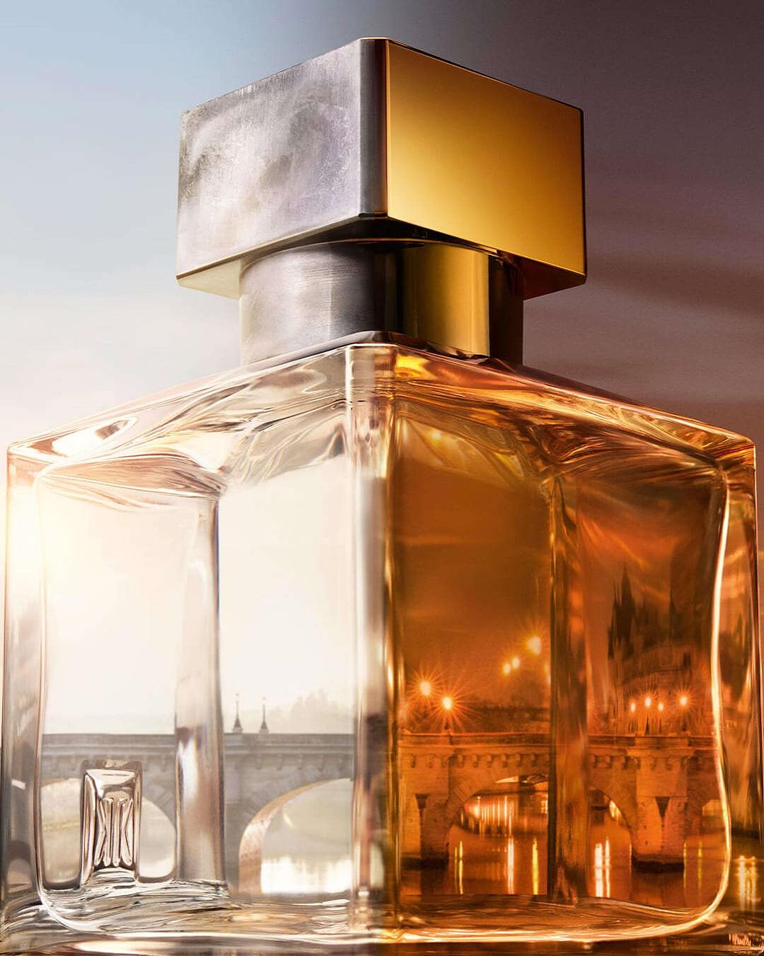 Close-up of a glass perfume bottle with a square shape and gold cap, reflecting a city bridge with lights in its surface.