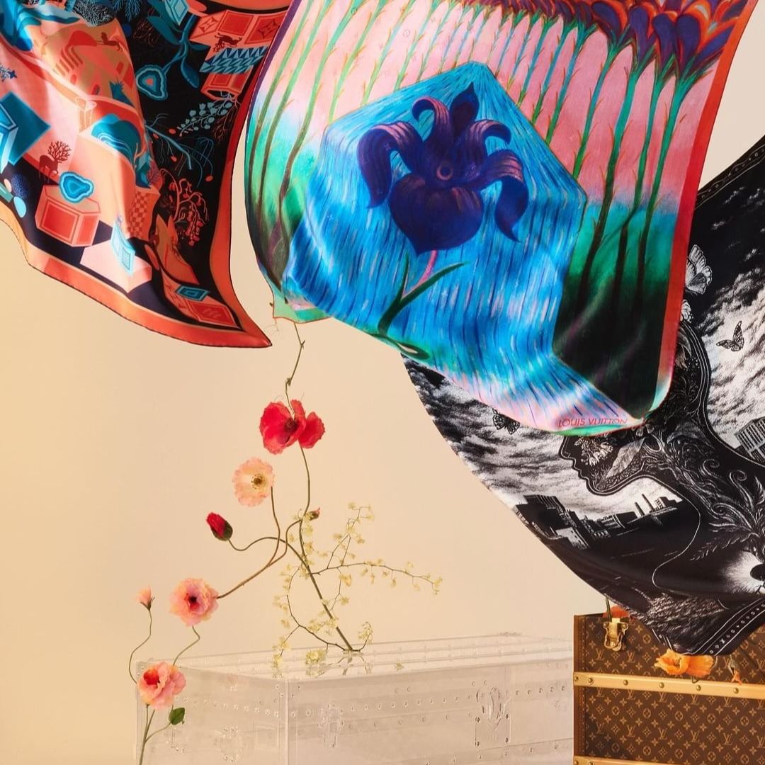 Three silk scarves with floral and graphic prints are suspended in the air with flowers, displayed with a vintage Louis Vuitton trunk.