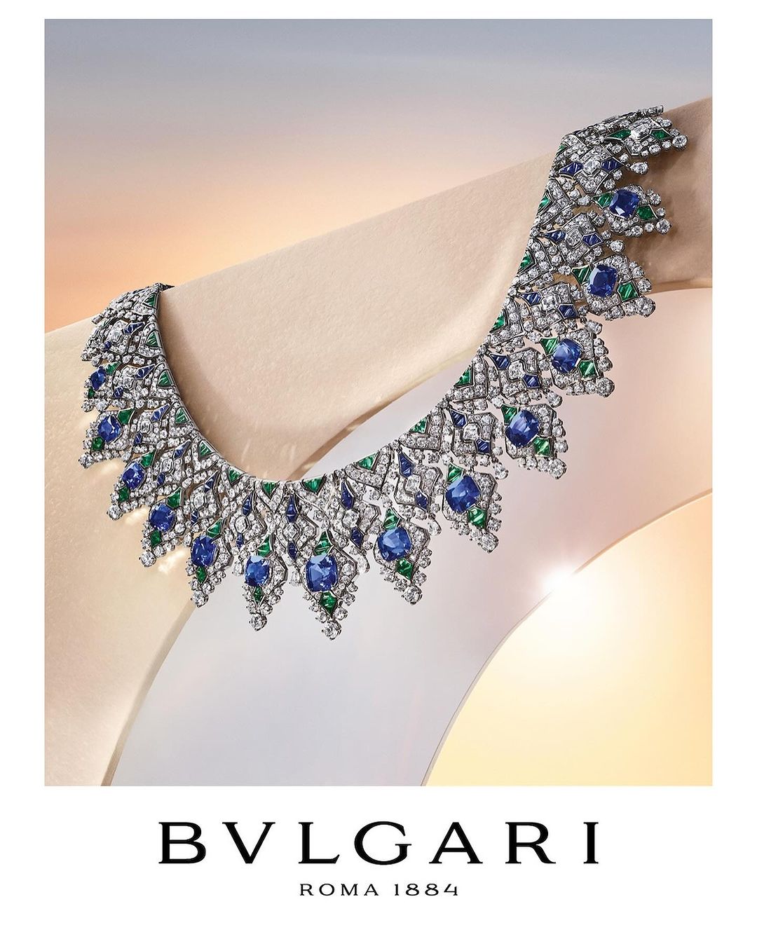 Close-up of a Bulgari necklace featuring blue and green gemstones and diamonds displayed on a cream-colored mannequin against a gradient backdrop.