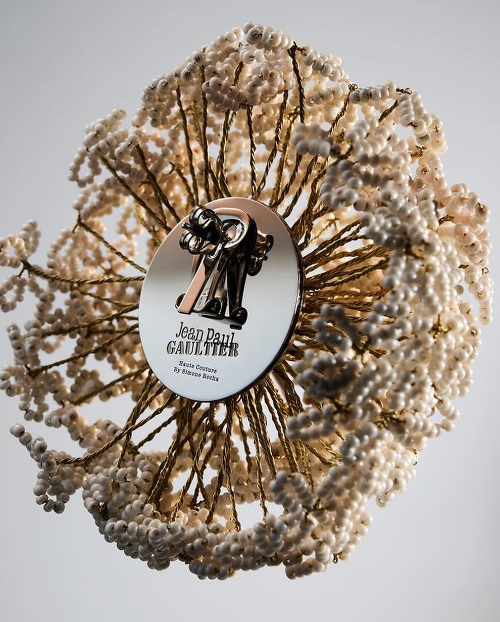 Close-up of beaded sculpture with logo of Jean Paul Gaultier, featuring intricate white beads, gold wire, and silver branding disk.