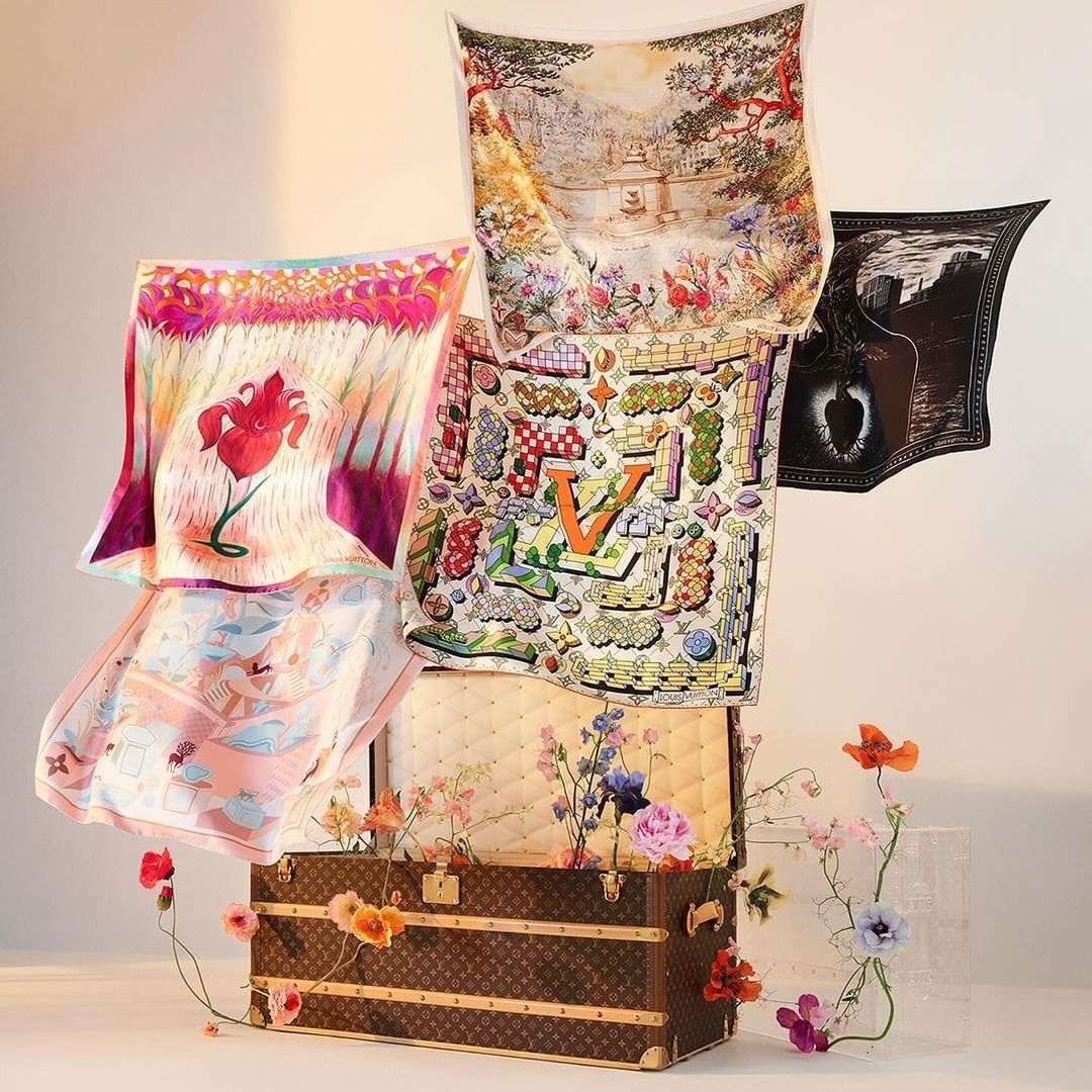 A collection of colorful silk scarves with floral and landscape designs arranged around a brown Louis Vuitton trunk with flowers.