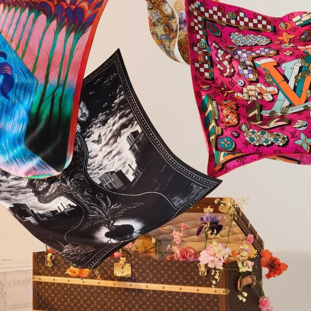 Colorful scarves arranged atop a Louis Vuitton trunk, complemented by flowers, against a cream backdrop, creating a striking still life.