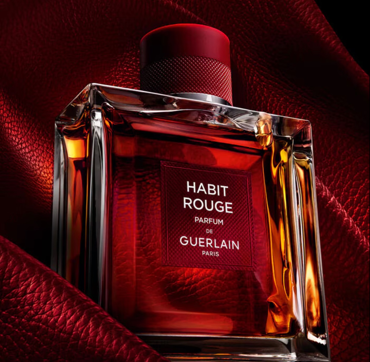 Guerlain Habit Rouge Parfum: a luxurious perfume bottle with a red cap and red liquid sits on a folded red leather surface in a dark studio.