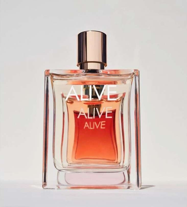Product shot of a square glass perfume bottle with warm orange liquid, gold accents, and the word 'ALIVE' printed on the front.