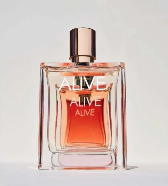 Product shot of a square glass perfume bottle with warm orange liquid, gold accents, and the word 'ALIVE' printed on the front.