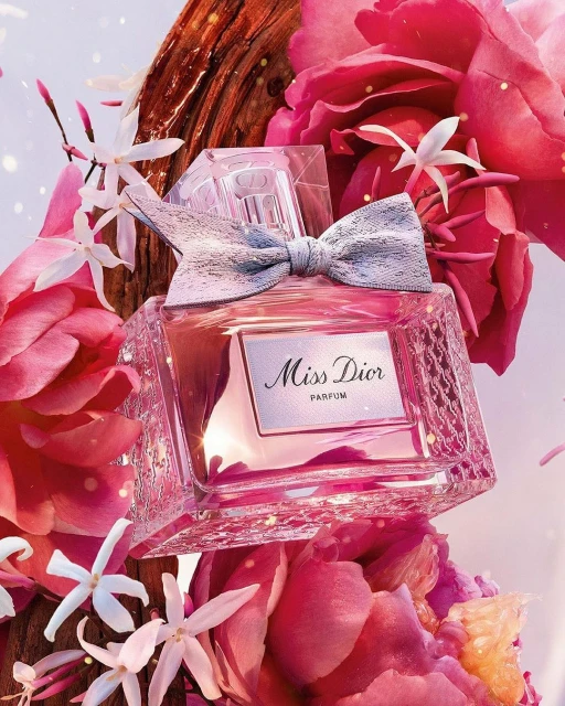 A close-up of a Miss Dior perfume bottle surrounded by pink and white flowers and a piece of wood, creating a floral and woody aesthetic.