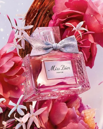 A close-up of a Miss Dior perfume bottle surrounded by pink and white flowers and a piece of wood, creating a floral and woody aesthetic.