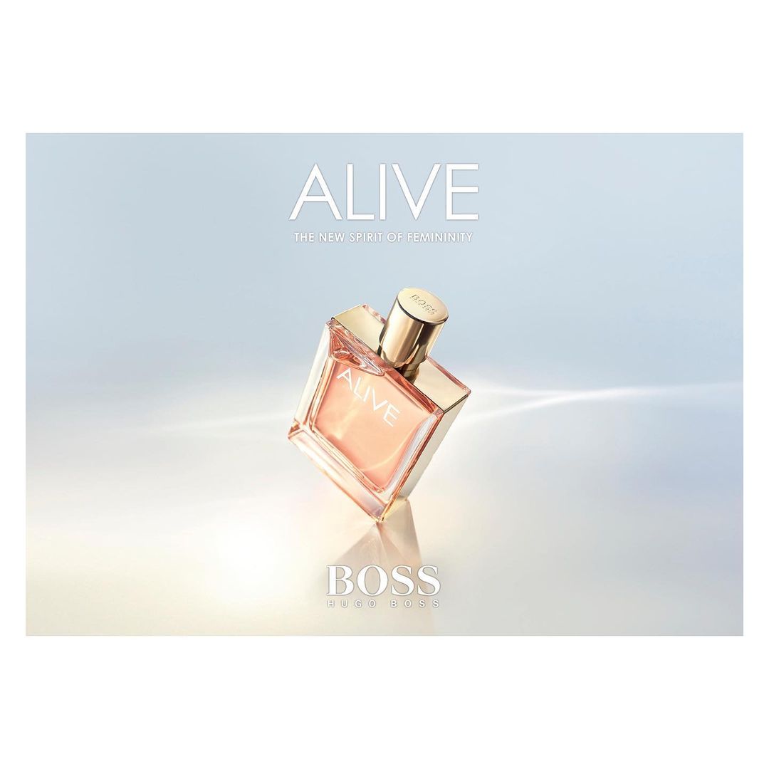 Hugo Boss Alive perfume bottle in square clear glass with metallic gold cap on a light blue and white gradient background.