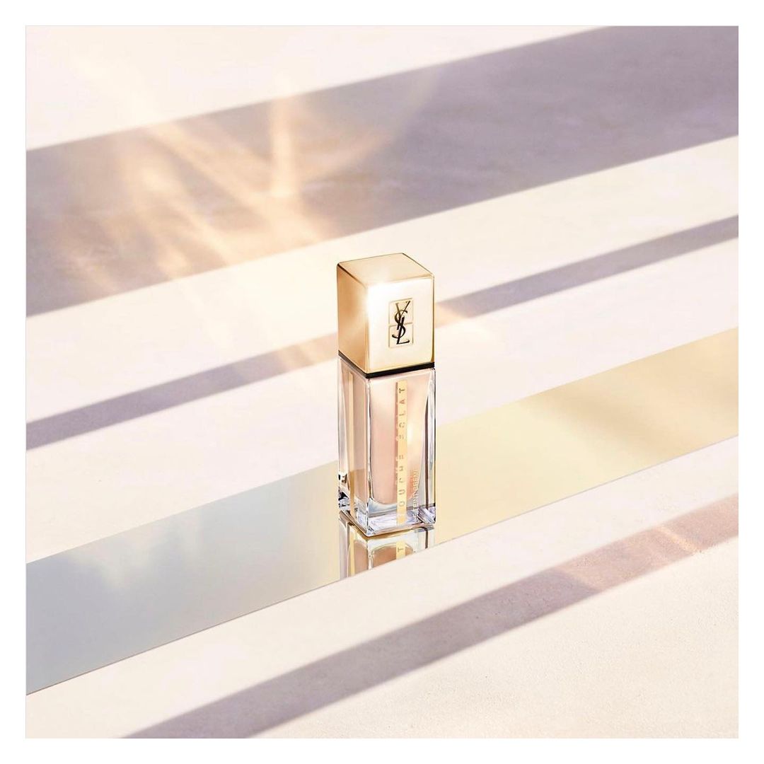 Elegant product shot of Yves Saint Laurent foundation with golden lid and clear bottle on a light surface with shadow stripes.