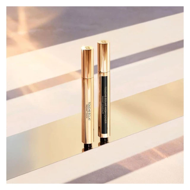Two golden YSL Touche Eclat concealers stand upright on a reflective gold surface against a softly lit beige background in studio shot.
