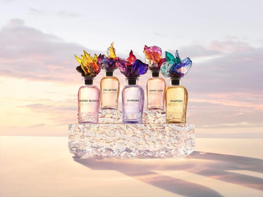 Five perfume bottles with colorful, abstract flower-shaped toppers are arranged on a shimmering, textured block against a soft sky backdrop.