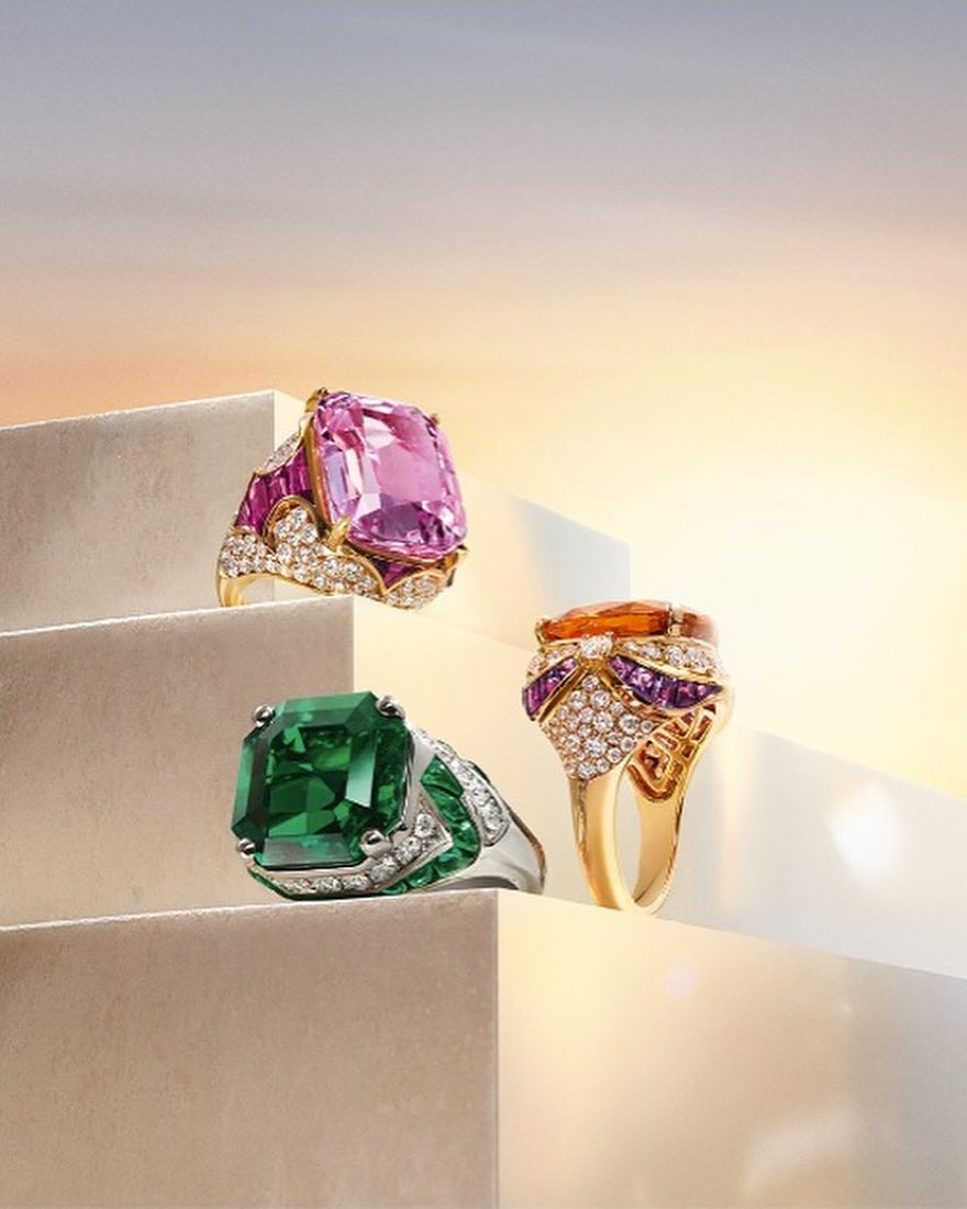 Three gemstone rings with diamonds displayed on ascending white steps against a soft gradient sky background in a studio shot.