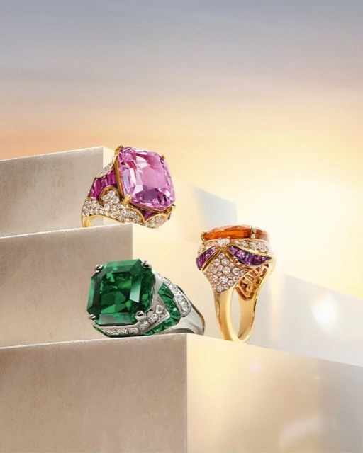 Three gemstone rings with diamonds displayed on ascending white steps against a soft gradient sky background in a studio shot.