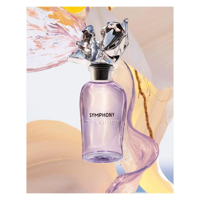 Louis Vuitton's Symphony perfume bottle in a studio shot with floral accents. The bottle is clear with a silver stopper. Fashion beauty shot.