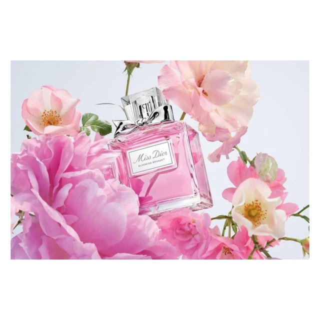 A bottle of Miss Dior Blooming Bouquet perfume is surrounded by pink and white roses against a soft, diffused white background.