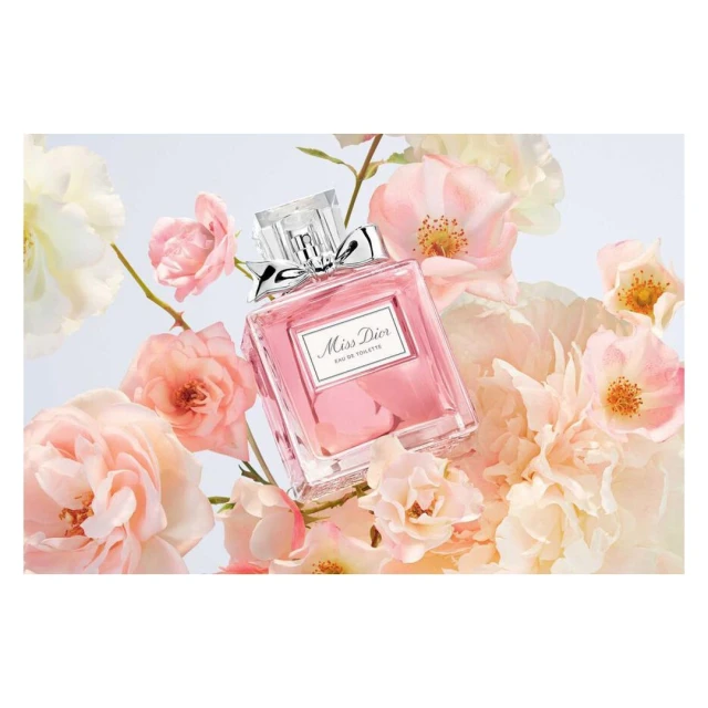 Close-up of a pink Miss Dior perfume bottle surrounded by pale pink and white roses on a light background, fashion, beauty, product photography.