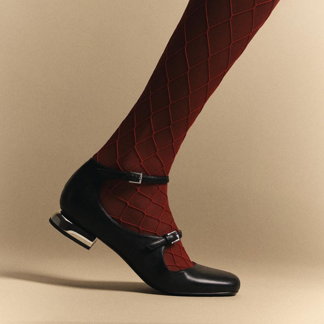 A close-up studio shot shows a woman's leg with foot in a black Mary Jane shoe with red patterned fishnet stockings against a beige backdrop.