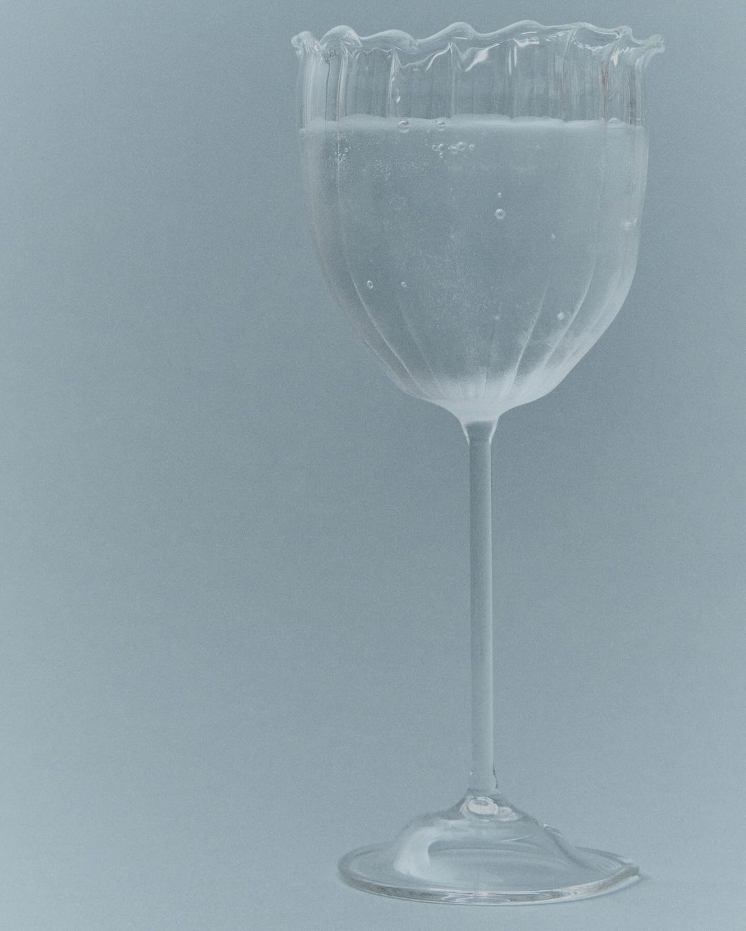 Elegant clear glass with a thin stem and scalloped rim filled with a clear liquid against a plain grey background.