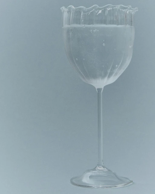 Elegant clear glass with a thin stem and scalloped rim filled with a clear liquid against a plain grey background.