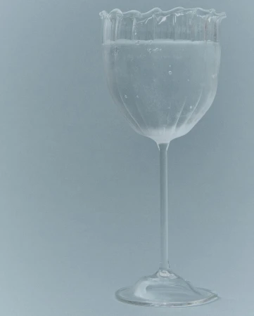 Elegant clear glass with a thin stem and scalloped rim filled with a clear liquid against a plain grey background.