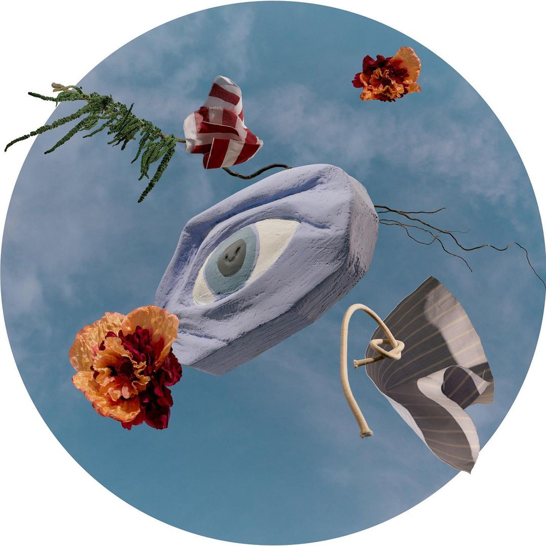 Surreal eye sculpture floats in a blue sky, surrounded by orange flowers and patterned fabric, creating a whimsical artistic composition.