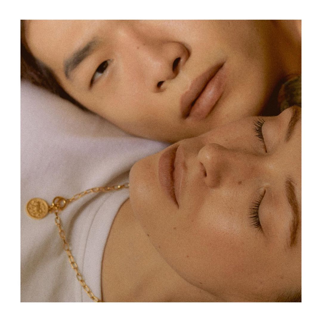 Close-up of a diverse couple with a light skin tone, lying together on a white surface, featuring a gold necklace and soft, neutral lighting.