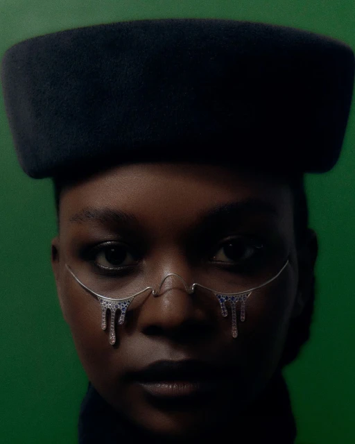 Close-up of a Black woman wearing a wide-brimmed hat and decorative eyewear with jewels, against a green backdrop in a studio setting.
