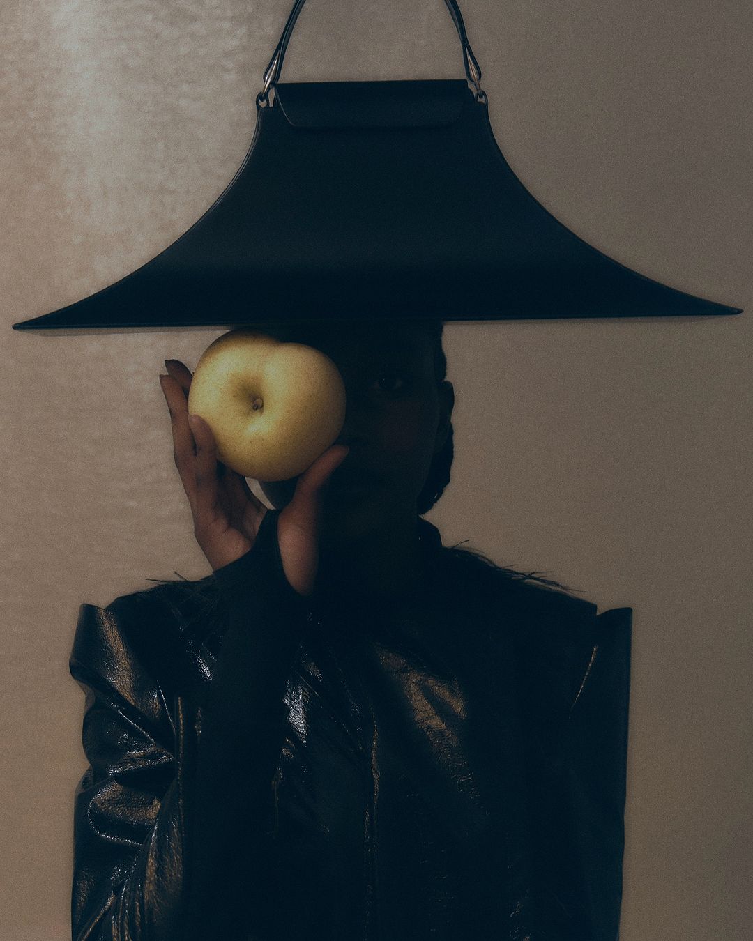 Woman in a black outfit holds a yellow apple in front of her face. Modern apron-style top frames the shot, indoor against a cream background.