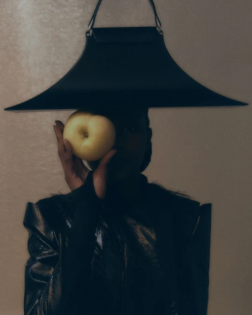 Woman in a black outfit holds a yellow apple in front of her face. Modern apron-style top frames the shot, indoor against a cream background.