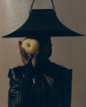 Woman in a black outfit holds a yellow apple in front of her face. Modern apron-style top frames the shot, indoor against a cream background.
